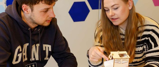 Ein junger Mann und eine junge Frau sitzen an einem Tisch, die Frau hält einen kleinen, selbstgebauten Roboter in der Hand und erklärt dem Mann etwas dazu.