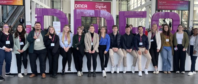 Eine Gruppe von 14 Studierenden und eine Dozentin stehen im Eingang einer Messehalle vor einer großen Skulptur aus lilanen Buchstaben: DMEA