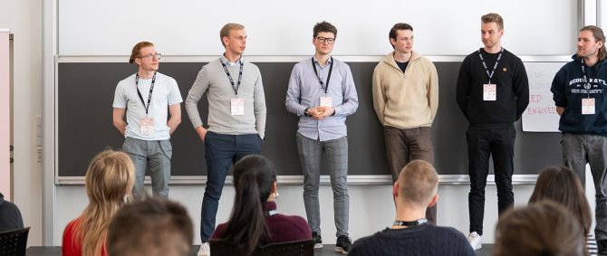 Eine Gruppe von Studierenden steht in einem großen Raum vor einem Publikum und hält einen Vortrag, neben ihnen stehen Roll-Ups von der SDU Case Competition.