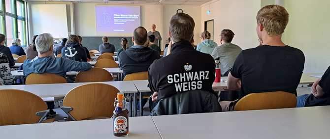 In einem Seminarraum sitzen viele Studierende, vorne steht ein Mann und hält einen Vortrag, seine Präsentation trägt den Titel "Ohne Wasser kein Bier!"