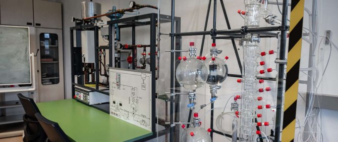 Das Bild zeigt ein regelungstechnisches Labor mit einer großen Glassapparatur zur Destillation. Daneben stehen ein grüner Labortisch, Stühle und ein Versuchstand mit Rohren und Messgeräten. Im Hintergrund sind Schränke und ein Abzug zu sehen.