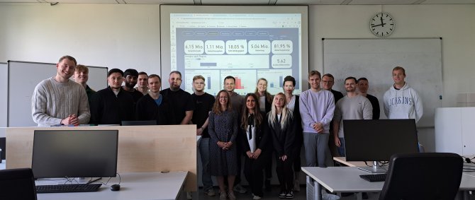 Eine Gruppe von ca. 30 Menschen steht in einem Seminarraum und lächelt in die Kamera, hinter ihnen an der Wand die Projektion einer Präsentationsfolie mit Diagrammen und anderen Daten.