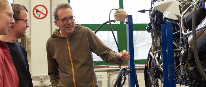 Zwei Studierende und ein Dozent stehen vor einem Motorrad.