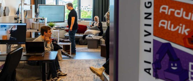 Blick durch eine offene Tür in ein Labor, das mit einem PC-Arbeitsplatz, einem Monitor, einem Regal und einem Sofa ausgestattet ist und in dem mehrere junge Menschen sitzen.