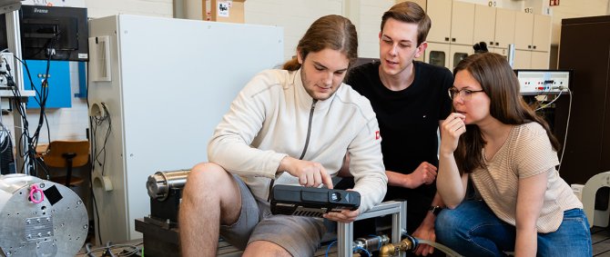 Zwei junge Männer und eine junge Frau knien in einem Labor zwischen technischen Apparaturen und schauen gemeinsam auf ein Messgerät.