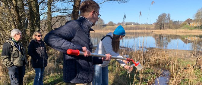 Zwei Menschen entnehmen eine Wasserprobe, im Hintergrund ist der See zu erkennen.