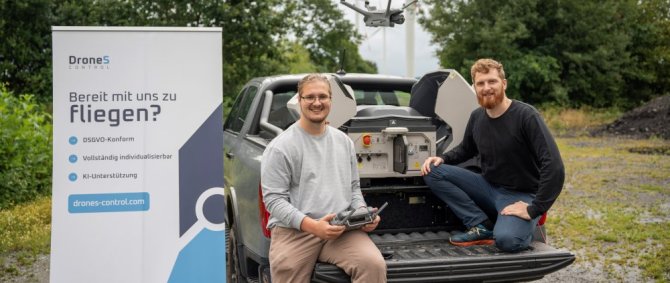 Zwei junge Männer hocken auf der Ladefläche eines Pichup-Trucks, der auf einem Feldweg steht. Einer steuert eine Drohne, neben ihm steht ein Roll-Up mit Text: DroneS Control. Bereit mit uns zu fliegen? DSGVO-konform, vollständig indiovidualisierbar, KI-Unterstützung. drones-control.com