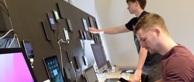 Zwei junge Männer an einem Arbeitsplatz mit verschiedenen mobilen Endgeräten. Die Tablets und Handys sind vor ihnen an einer Wand angebracht.