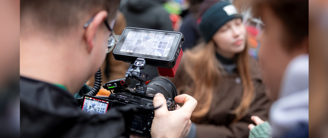 Nahaufnahme über die Schulter eines jungen Mannes, der mit einer Kamera eine andere Studentin filmt.