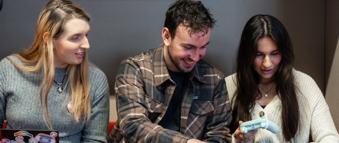 Drei Studierende sitzen in einer Sitzecke und arbeiten gemeinsam an Laptops, zwei haben Spielekonsolen-Controller in der Hand.