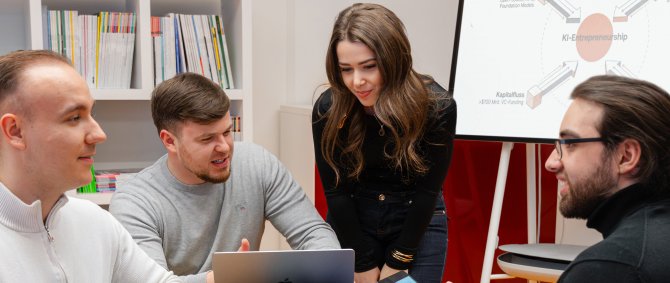 Vier Studierende sitzen um einen Tisch und diskutieren, hinter ihnen ein großer Monitor mit einem Schaubild zu KI-Entrepreneurship.