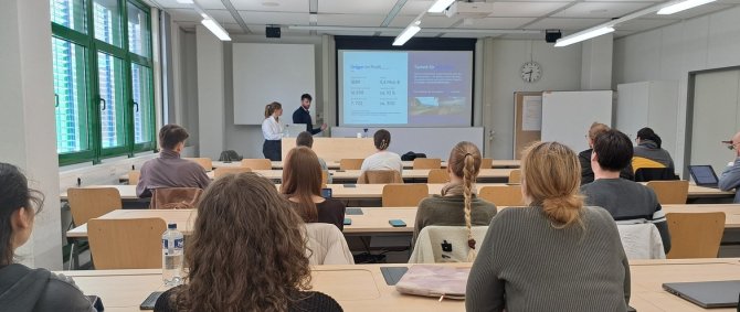 In einem Seminarraum sitzen Studierende und lauschen einem Vortrag, den eine junge Frau und ein junger Mann vorne halten.