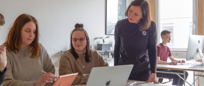 Studierende arbeiten in einem hellen, freundlichen Raum an Laptops. Eine DSozentin steht daneben und hört interessiert zu.