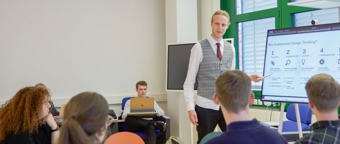Seminarszene: Student steht neben Dozent an einem Bildschirm und erläutert etwas. Im Vordergrund andere Studierende um einen Tisch.