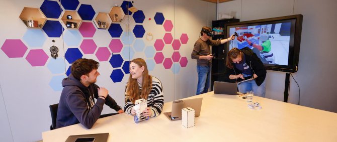 In einem Seminarraum mit bunten Kacheln an der Wand sitzen zwei Studierende an einem Tisch und unterhalten sich, auf dem Tisch vor ihnen steht ein kleiner Roboter. Im Hintergrund steht ein Dozent vor einem Monitor und trägt eine VR-Brille, neben ihm eine weitere Studentin, die etwas auf einem Tablet nachschaut.