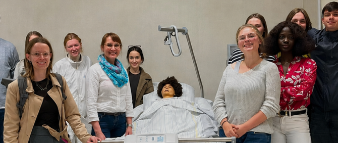 Eine Gruppe von etwa 12 Studierenden steht um ein Pflegebett mit einer Puppe drumherum und lächelt fröhlich in die Kamera.