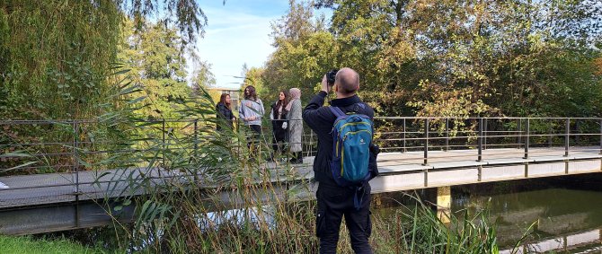 Ein junger Fotograf steht auf einer Wiese und fotografiert vier Studierende, die auf einer Brücke posieren.