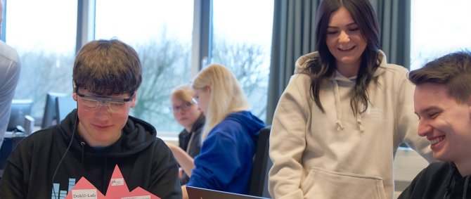 Studierende sitzen, einer trägt eine Eye Tracking-Brille. Hinter ihnen stehen Lehrende. Sie lachen.