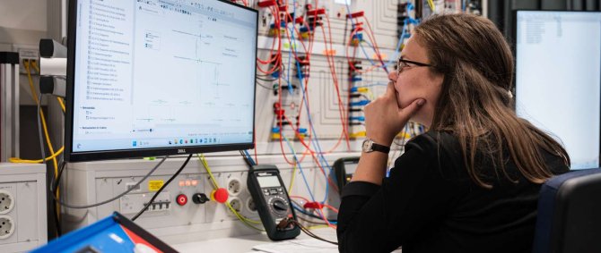 Eine junge Frau sitzt an einem PC-Arbeitsplatz mit einer großen elektrischen Schaltwand und blickt konzentriert auf den Bildschirm.