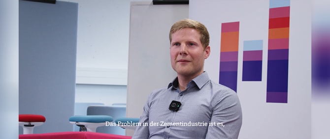 Ein junger Mann im hellblauen Hemd sitzt vor einem Roll-Up mit dem Logo der Hochschule Flensburg und spricht in die Kamera.