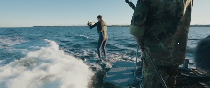 Ein junger Mann im Neoprenanzug mit Filmkamera springt vom Heck eines fahrenden Marine-Schiffes.