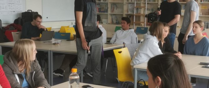 In einem Seminarraum sitzen Studierende an Gruppentischen, einige mit Laptops vor sich, andere in Diskussionen vertieft. Zwischen den Tischen stehen andere Studierende mit Kameraausrüstung.