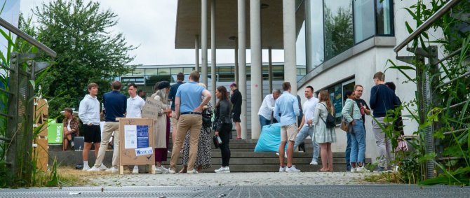 Mehrere Grüppchen von Menschen verschiedenen Alters stehen neben einem modernen Gebäude mit Säulen auf und an einer Treppe und unterhalten sich angeregt.