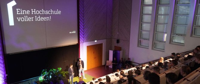 In einem gut gefüllten Hörsaal stehen zwei Männer vorne im Scheinwerferlicht und sprechen zum Publikum, über ihnen eine Projektion mit Text: Eine Hochschule voller Ideen!