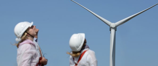 Vorn im Bild sind unscharf zwei junge Frauen in weißen Arbeitsanzügen und mit Helm zu erkennen, sie schauen schräg nach oben auf die Windenergieanlage, die scharf zu erkennen ist. Der Bildhintergrund ist der blaue Himmel.