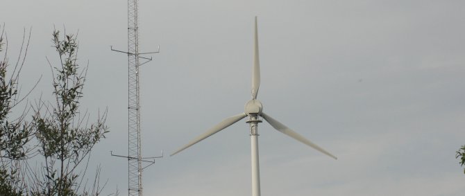 Windenergieanlage und dahinter Windmast vor bewölktem Himmel. Links vorn im Bild ist Teil eines Baumes zu erkennen.