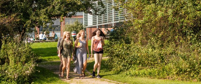 Drei Studentinnen laufen an einem sommertag über den Campus. Sie sind von Grün, Büschen und Rasen umgeben, im Hintergrund ist gerade noch so ein Ggebäude und weitere Menschen an Tischen zu erkennen. Die jungen Frauen lachen und tragen sommerliche Kleidung.