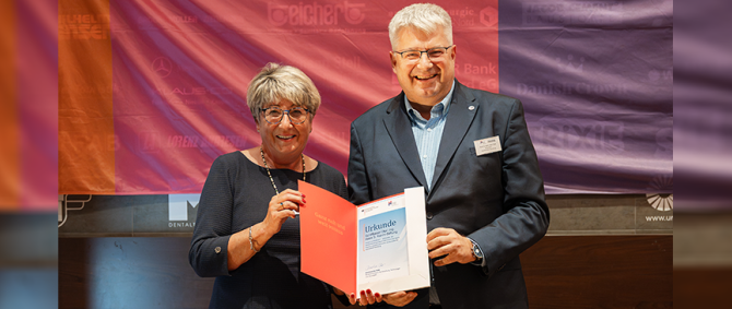 Eine Frau und ein Mann, beide fortgeschrittenen Alters und in Anzug bzw. Kostüm, stehen vor einer bunten Flagge und halten lächelnd eine Urkunde in die Kamera.