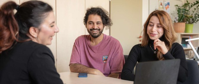 Ein junger Mann und eine junge Frau sitzen an einem Schreibtisch einer Frau im schwarzen Blazer gegenüber; alle drei schauen auf einen Laptopbildschirm und lachen fröhlich.