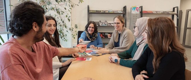 Um einen Tisch sitzt eine Gruppe von internationalen Studierenden und Frauen im Gespräch, eine von ihnen reicht einem Studenten einen Flyer über den Tisch.