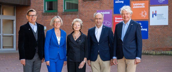 Drei Männer und zwei Frauen posieren vor einem Hochschulgebäude für ein Gruppenbild. Im Hintergrund u.a. Kacheln in Hochcshulfarben und das Hochschul-Logo.