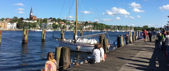 Der Flensburger Hafen im strahlenden Sonnenschein. Auf dem Bohlwerk, einer hölzernen Anlegebrücke, sitzen Menschen am Wasser, ein hölzernes Segelschiff liegt vor Anker.