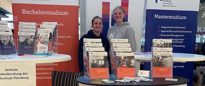 Zwei junge Frauen stehen an einem Messestand der Hochschule Flensburg mit Rollups und Flyern und lächeln in die Kamera.