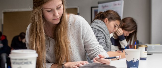 Studentin mit Stift in fder Hand an einem Tisch sitzend, auf dem Tisch Kaffeebecjer und im Hintergrund zwei weitere Studentinnen, die über Unterlagen gebeugt sind und Stifte in den Händen halten.