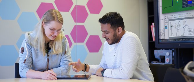 Eine junge blonde Frau und ein junger Mann mit schwarzen Haaren und Bart sitzen an einem Tisch, unterhalten sich und haben ein Tablet zwischen sich liegen. Im Hintergrund ein großer Monitor mit einer technischen Zeichnung eines Windrads und eine Wand mit bunten Hexagons.