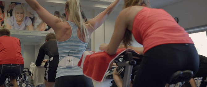 Junge Menschen auf Spinning-Fahrrädern und eine Trainerin, die motivierend die Arme hochstreckt.