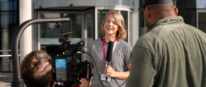 Eine junge Frau steht vor einem modernen Vorlesungsgebäude mit Glasfront, dem Flensburger audimax. Sie spricht in ein Mikrofon, während zwei junge Männer sie mit einer Kamera filmen.