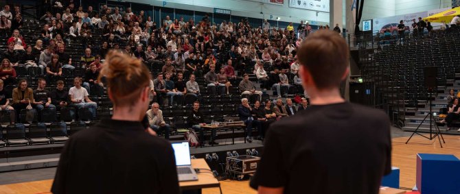 Zwei junge Männer in schwarzen T-Shirts von hinten fotografiert - sie stehen auf einer Bühne mit Blick auf eine gut gefüllte Tribüne, vor ihnen sind auf der Bühne bunte Sitzwürfel verteilt.