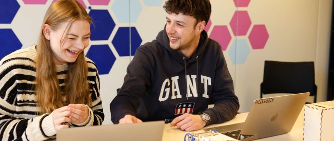 Eine Studentin und ein Student sitzen an einem Tisch, vor sich aufgeklappte Laptops, und lachen herzlich. An der Wand hinter ihnen sind rosane und blaue Kacheln.
