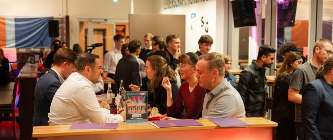 Bild einer Feier: Mittig sitzen mehrere Menschen um einen Tisch, essen und reden miteinander. Rechts stehen Menschen in einer Schlange.