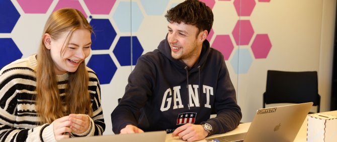 Eine Studentin und ein Student sitzen an einem Tisch, vor sich aufgeklappte Laptops, und lachen herzlich. An der Wand hinter ihnen sind rosane und blaue Kacheln.