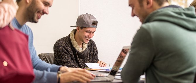 Drei Studierende sitzen an einem Tisch und arbeiten fröhlich an ihren Laptops.
