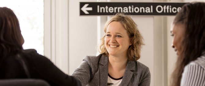Staff member of the International Office talking to two students who can be seen from the back and slightly blurry. There's a sign saying "International Office" at the top of the picture.