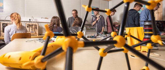 Molekülähnliches Model im Vordergrund, dahinter ein Professor und mehrere Studierende im Gespräch und an der Arbeit an 3D-Druckern.