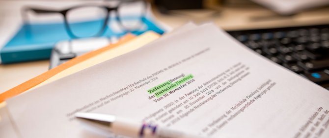 Ausdruck einer Satzung, der Titel ist grün markiert, im Hintergrund sind eine Brille und eine Tastatur zu erkennen, vorne ein Stift mit Hochschullogo