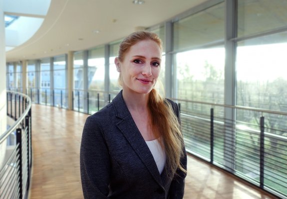 Porträtfoto: Lena Marie Beck im audimax. Sie trägt einen dunklen Blazer über einem hellen Oberteil. Ihre langen dunkelblonden Haare liegen in einem Zopf über ihrer linken Schulter. Hinter ihr ist die Fensterfront des audimax mit Ausblick auf den grünen Campus und der Parkettboden zu sehen.